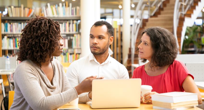 Two women and man sit at table with computer in library talking.