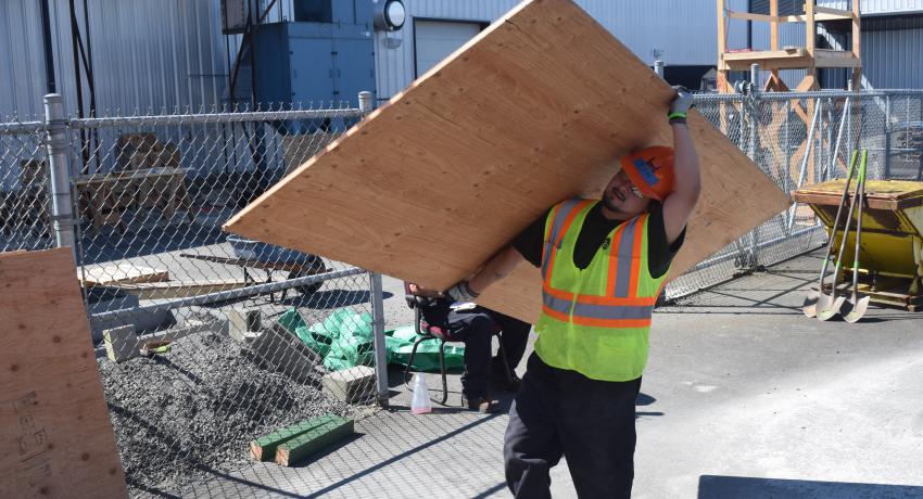 Young man carries large piece of sheet wood on construction site. 