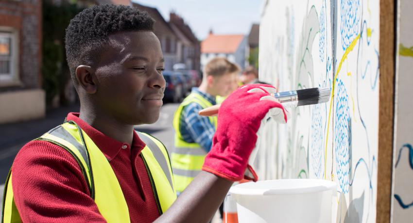 A young man helps to paint a mural in a public space. 