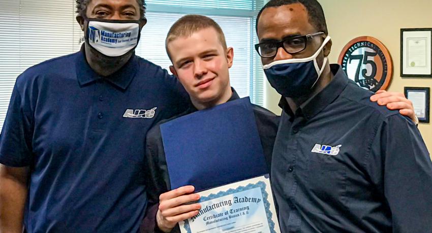 A graduating youth stands with his Manufacturing Academy teachers holding his certificate of completion. 