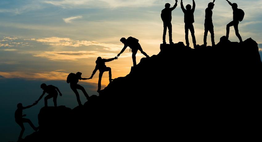 A group of silhouetted people help each other reach a mountain top.  
