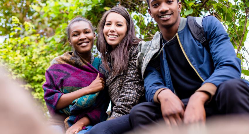 Three youth sit together in a park smiling. 