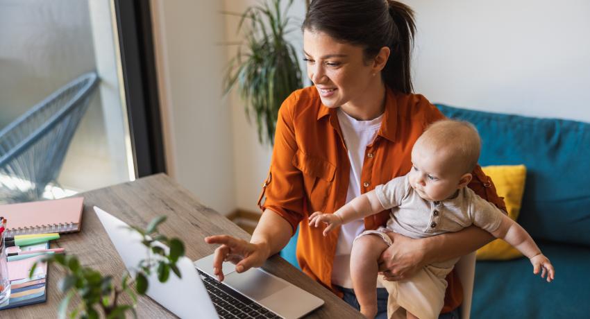 Young mother with dark hair carries baby in one arm while navigating laptop with her right hand.