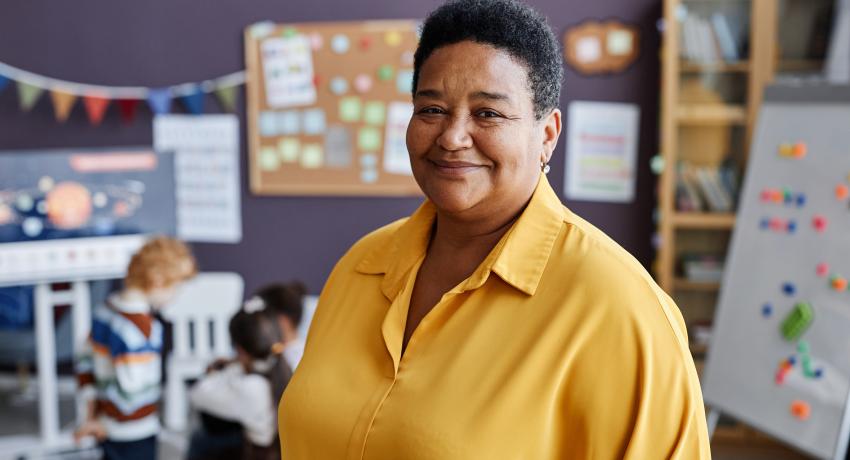 Woman of color wearing yellow blouse and standing proud in a child care classroom.