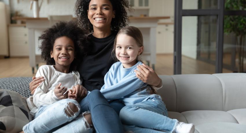 Portrait of smiling African American mother hugging two little daughters, multiracial family concept, sitting on cozy couch in living room, posing for photo, looking at camera, adopted child