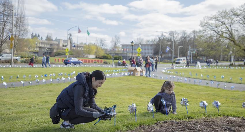Woman and girl kneel down on grass and plant silver and blue pinwheels. Pinwheels in the ground are also visible in the background.