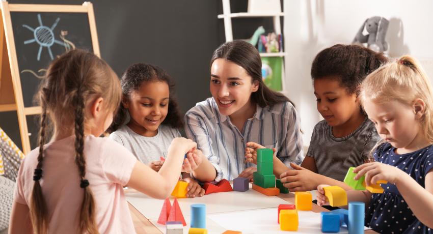 Woman with long, dark hair sits at desk engaging with four young girls in child care classroom.